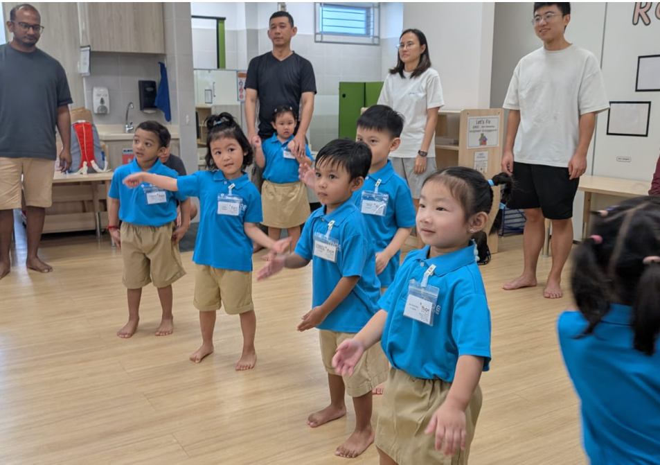 Young children in blue uniforms stand barefoot in a classroom, with three adults watching behind.